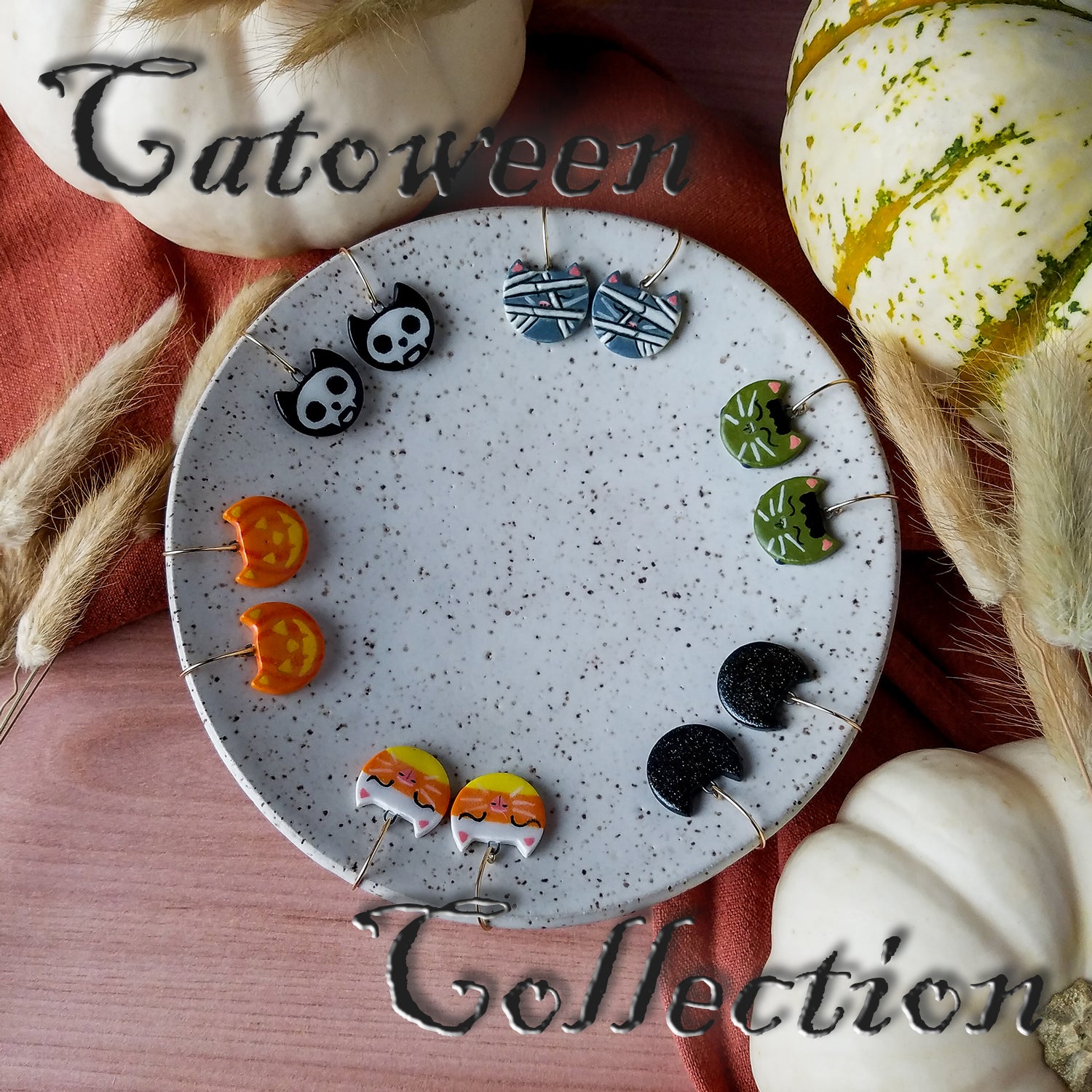 Cat-head-shaped earrings from the Catoween Collection surround the inside edge of a speckled white porcelain dish on a wooden table with an orange cloth, white pumpkins, and dried grasses as accents.  Earring designs include each of the one-of-a-kind Catoween Collection pieces.  Clockwise from upper left: Skelecats, Meowmies, Catenstein’s Meownsters, Black Magic Kittens, Catty Corn, and Cat-O-Lanterns.  Spooky text overlay says “Catoween Collection.”