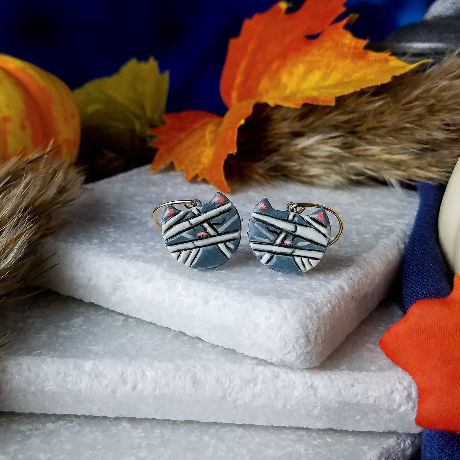 Porcelain cat heads sculpted and painted as Meowmies/Mummies complete with wrappings and happy features on gold-filled curved ear wires.  Earrings are on a stack of white marble blocks, surrounded by Autumnal items – leaves, pumpkins, and dried grasses – with a blue night sky background.