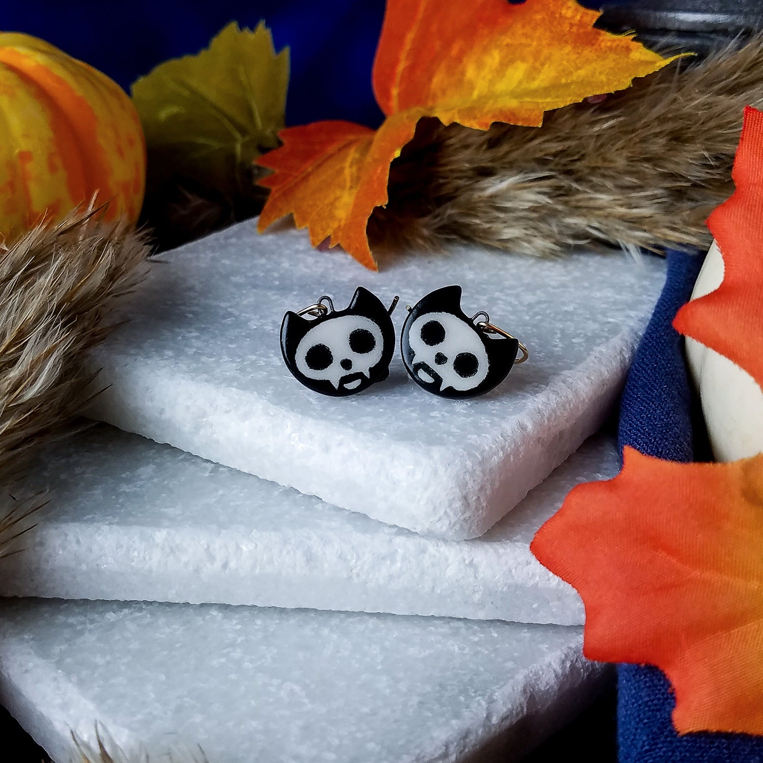 Porcelain cat heads painted as Skelecat cat skulls on gold-filled curved ear wires.  Earrings are on a stack of white marble blocks, surrounded by Autumnal items – leaves, pumpkins, and dried grasses – with a blue night sky background.