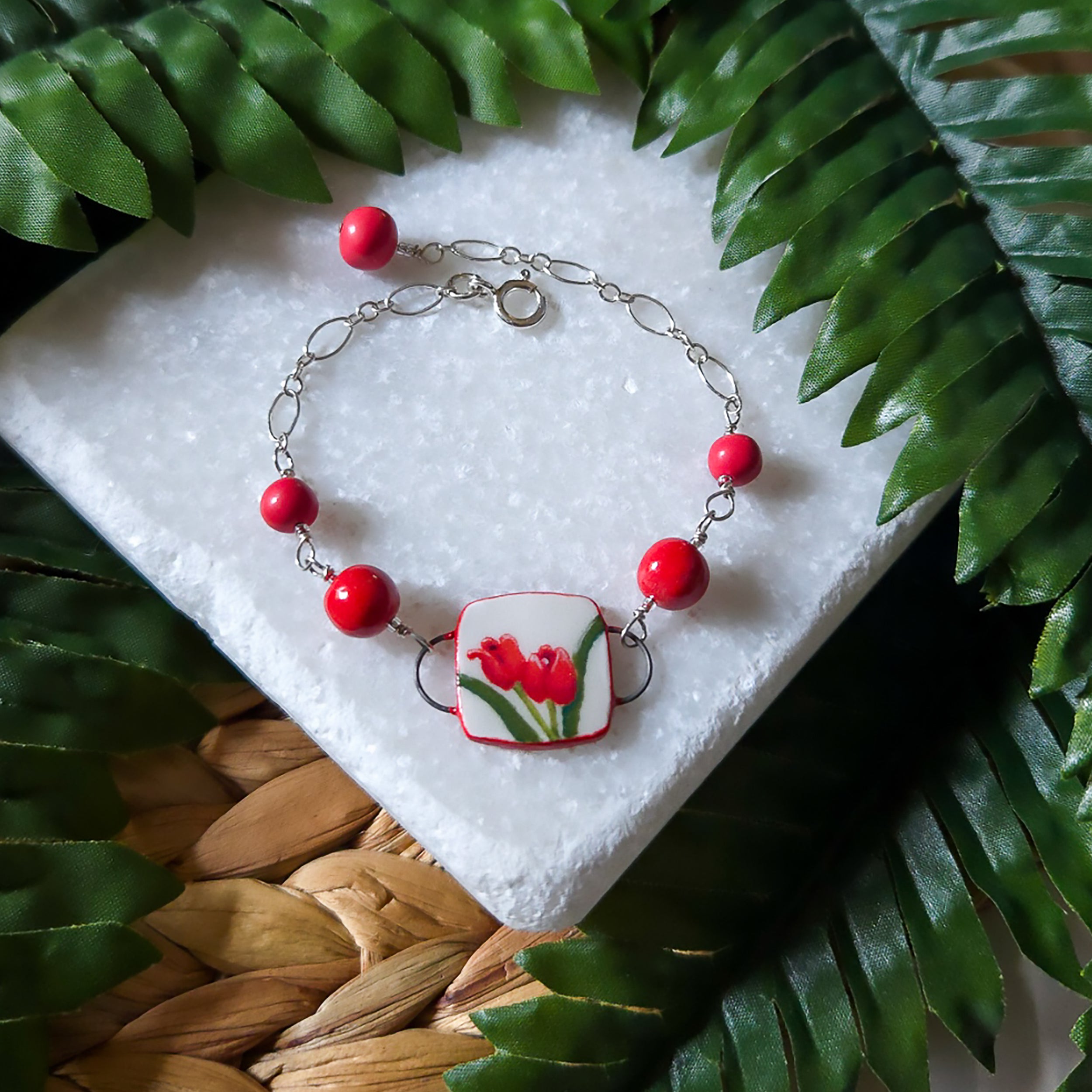 A bracelet with 2 red tulips on the focal panel rests on a stone slab surrounded by greenery.