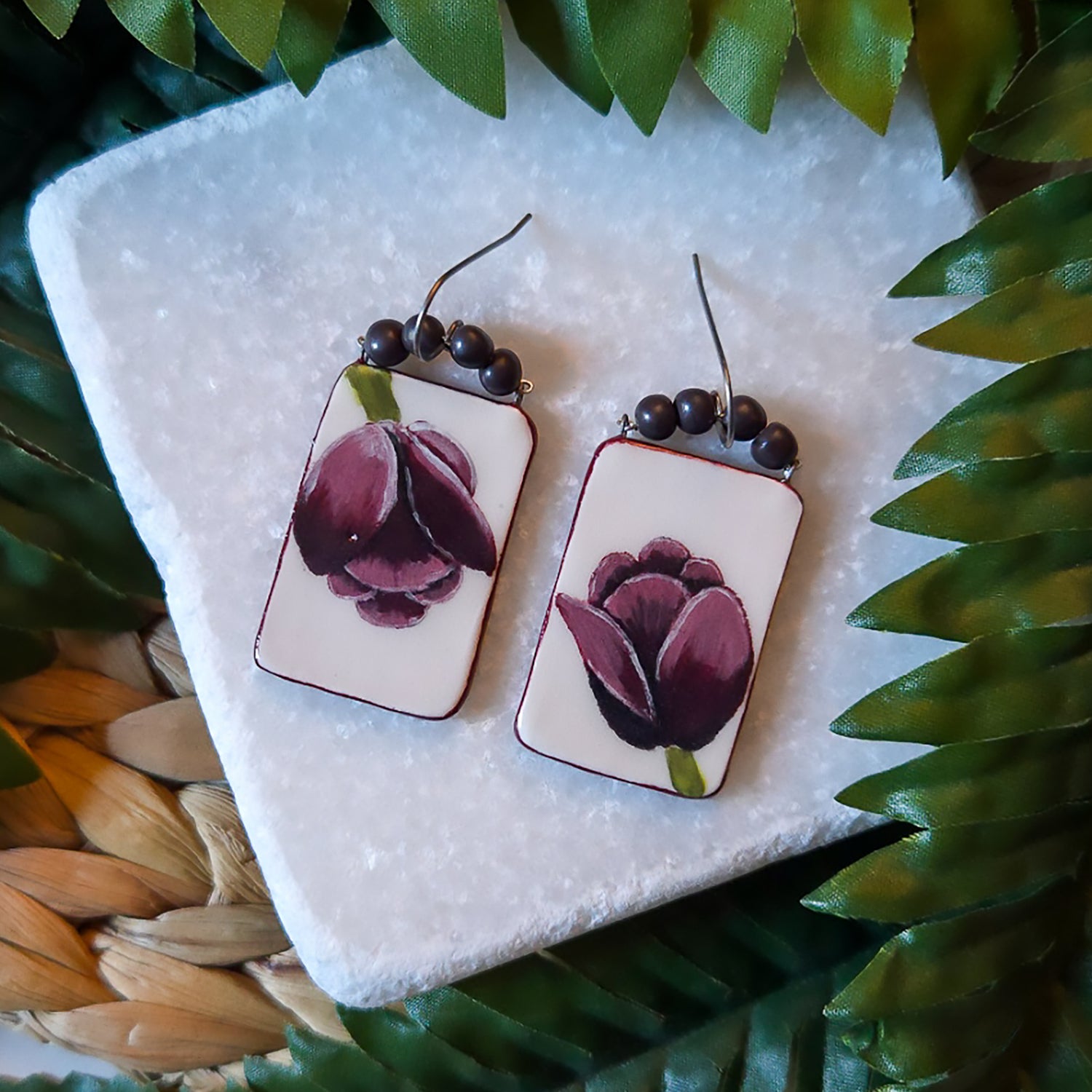 The pair of Tulip Panel Dangle Earrings in Burgundy rests on a stone slab surrounded by greenery.  One tulip faces up, the other down, so they seem to be looking at each other.